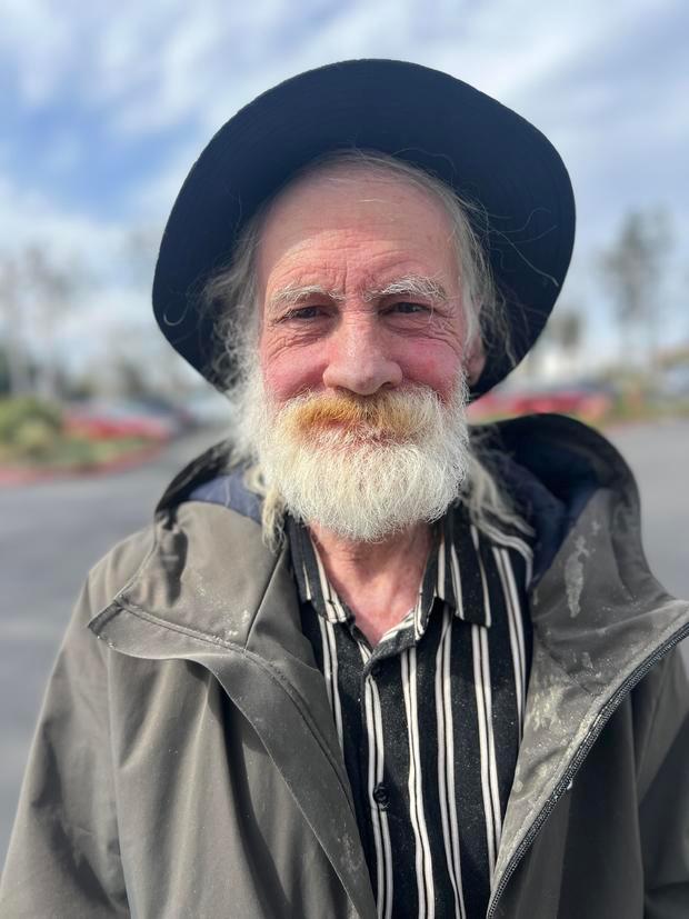 Cashman Whiteley, wearing a black and white striped shirt, green jacket, and black hat, smiles at the camera