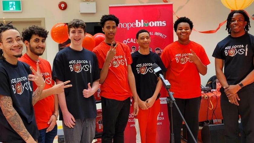 A group of seven boys are standing in front of a banner and table, smiling at the camera. They are wearing shirts that say "Hot Cocoa Boys" and in front of them is a mic.