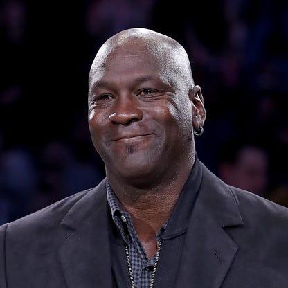 Michael Jordan, wearing a black shirt and blue jeans, kneels next to a young boy. The boy is wearing a black and white jacket that says "Jordan" on it and blue jean shorts, and a black baseball cap.