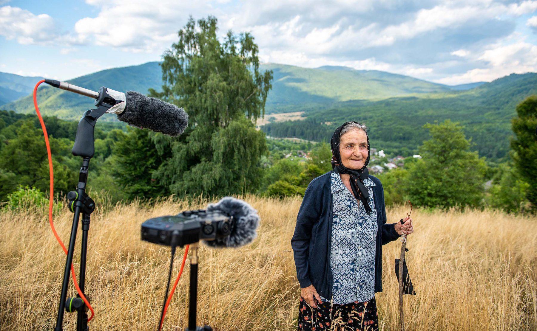 woman standing in a field with mountains in the background and recording equipment in front of her