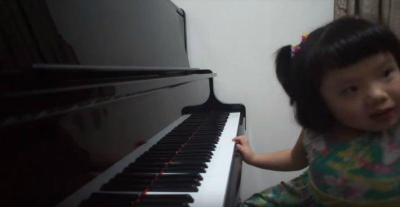 A little girl with short black hair, wearing a green dress printed with pink and yellow flowers, sits on the left side of the image in front of a piano.