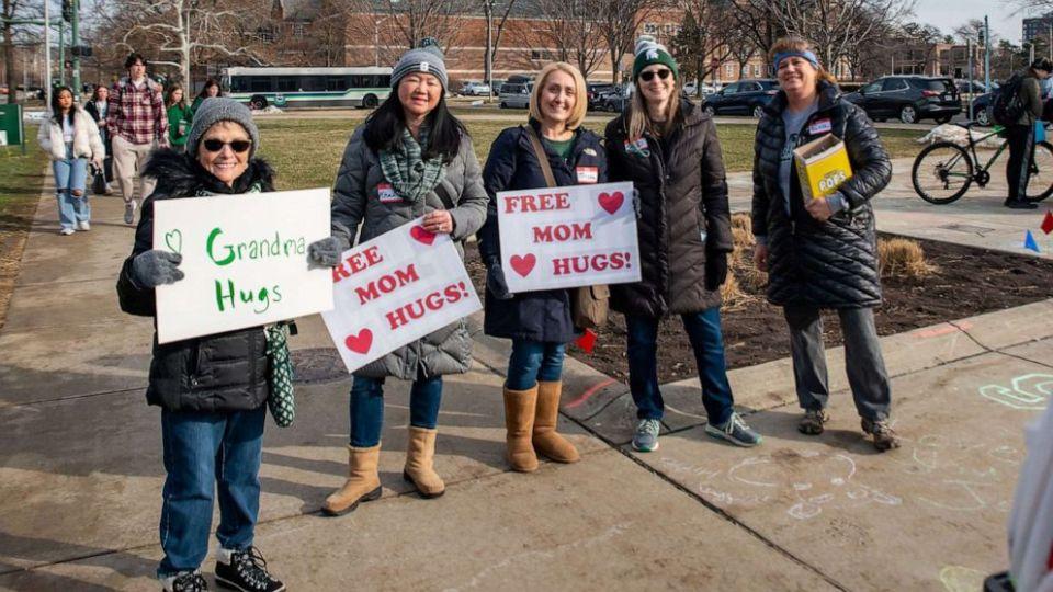 women are wearing winter clothes, standing outside on a sidewalk, holding signs that say, free mom hugs.