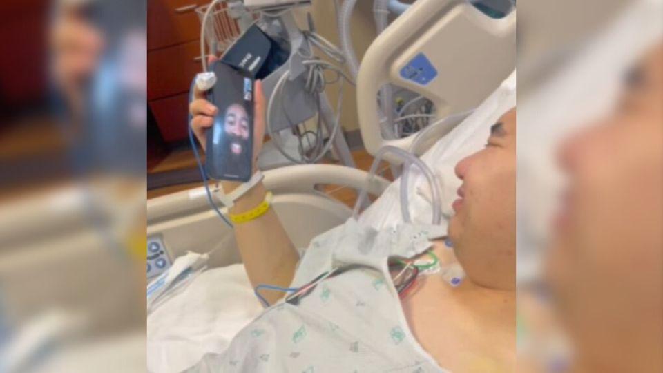 a man lies in a hospital bed smiling, while holding a phone. he is video chatting with another person who is smiling.