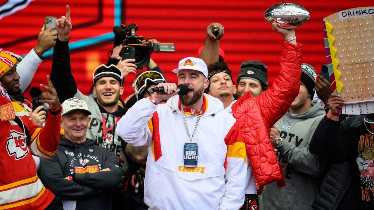 Kansas City Chiefs tight end Travis Kelce hypes up the crowd at the Super Bowl victory celebration rally. He is standing in between teammates, wearing a white sweatshirt, speaking into a mic.