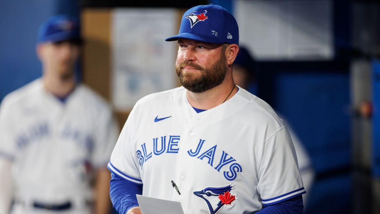 John Schneider, the manager for the Toronto Blue Jays, is wearing a baseball cap and shirt with the team logo, standing in the locker room.