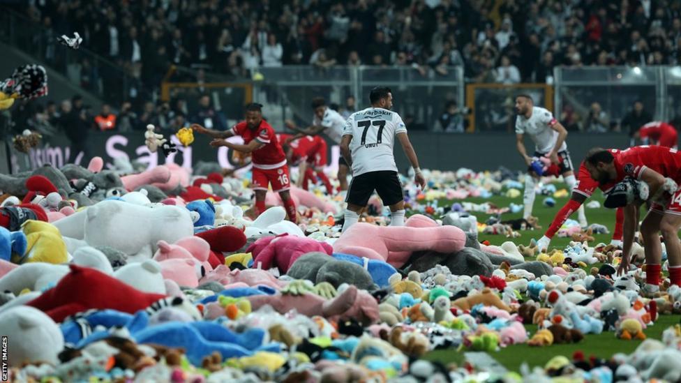 Stuffed toys cover the ground of a soccer field, as soccer players wearing red and white jerseys move the toys to the side.